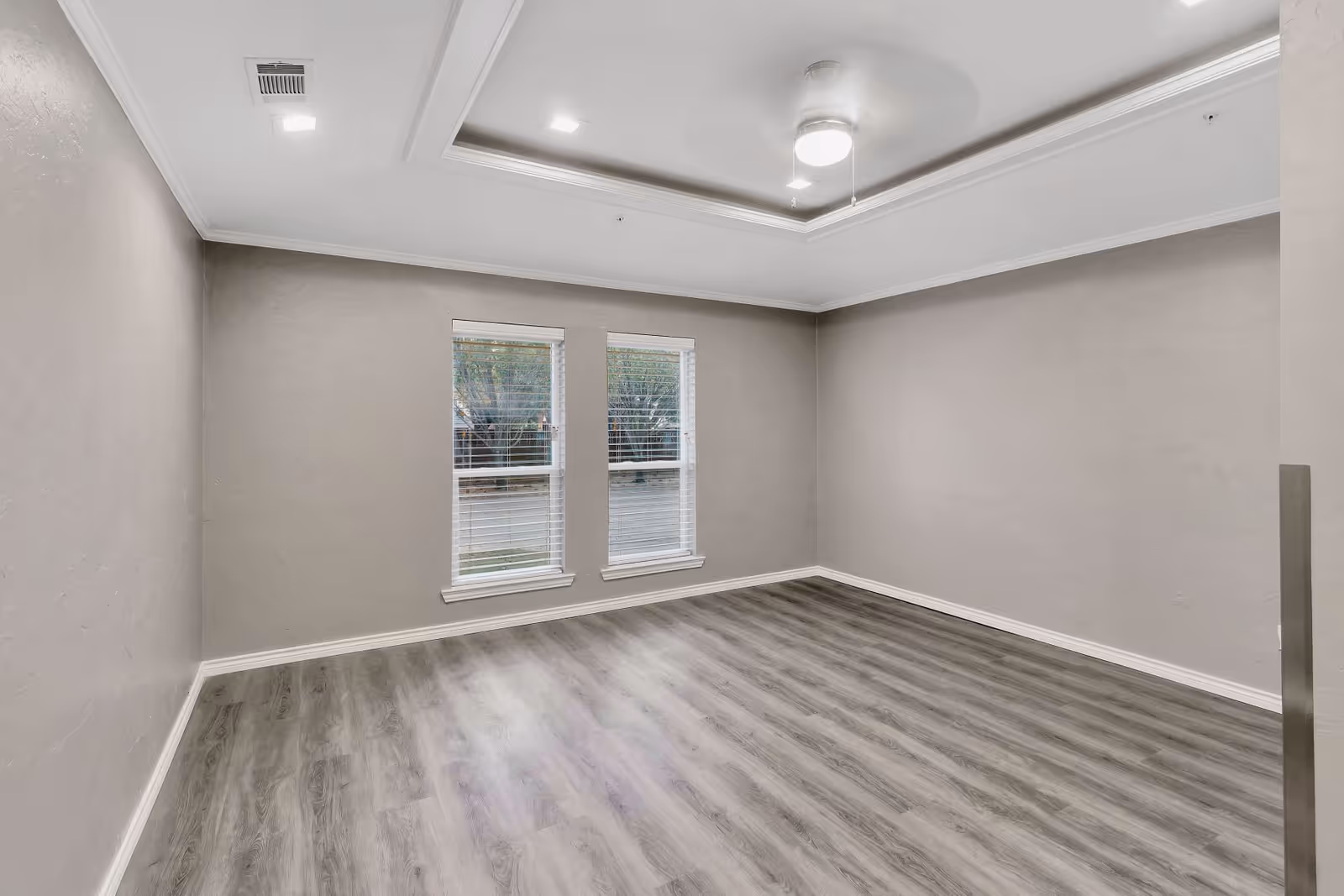 Empty room with gray walls, wood-look flooring, two windows, and a tray ceiling with a ceiling fan light.