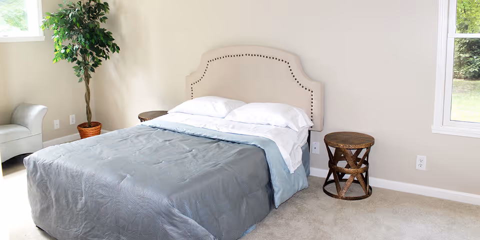 A bedroom with a neatly made bed featuring a beige upholstered headboard and gray bedding. There are two round wicker side tables on either side of the bed, a small gray armchair in the corner, a potted plant, and a window showing greenery outside.