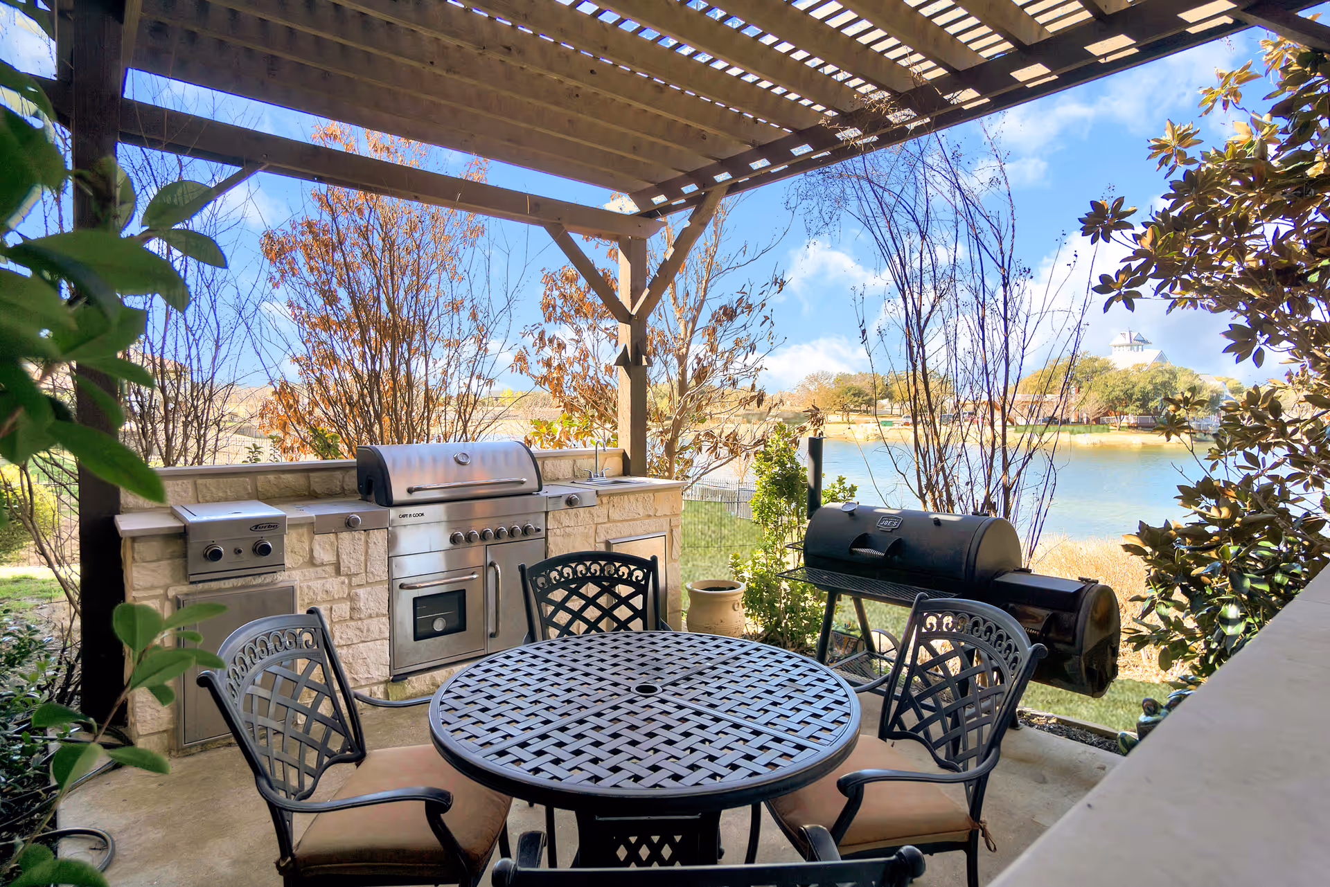 Covered outdoor patio with a round metal table and chairs, a built-in stainless grill and a smoker overlooking a lake.