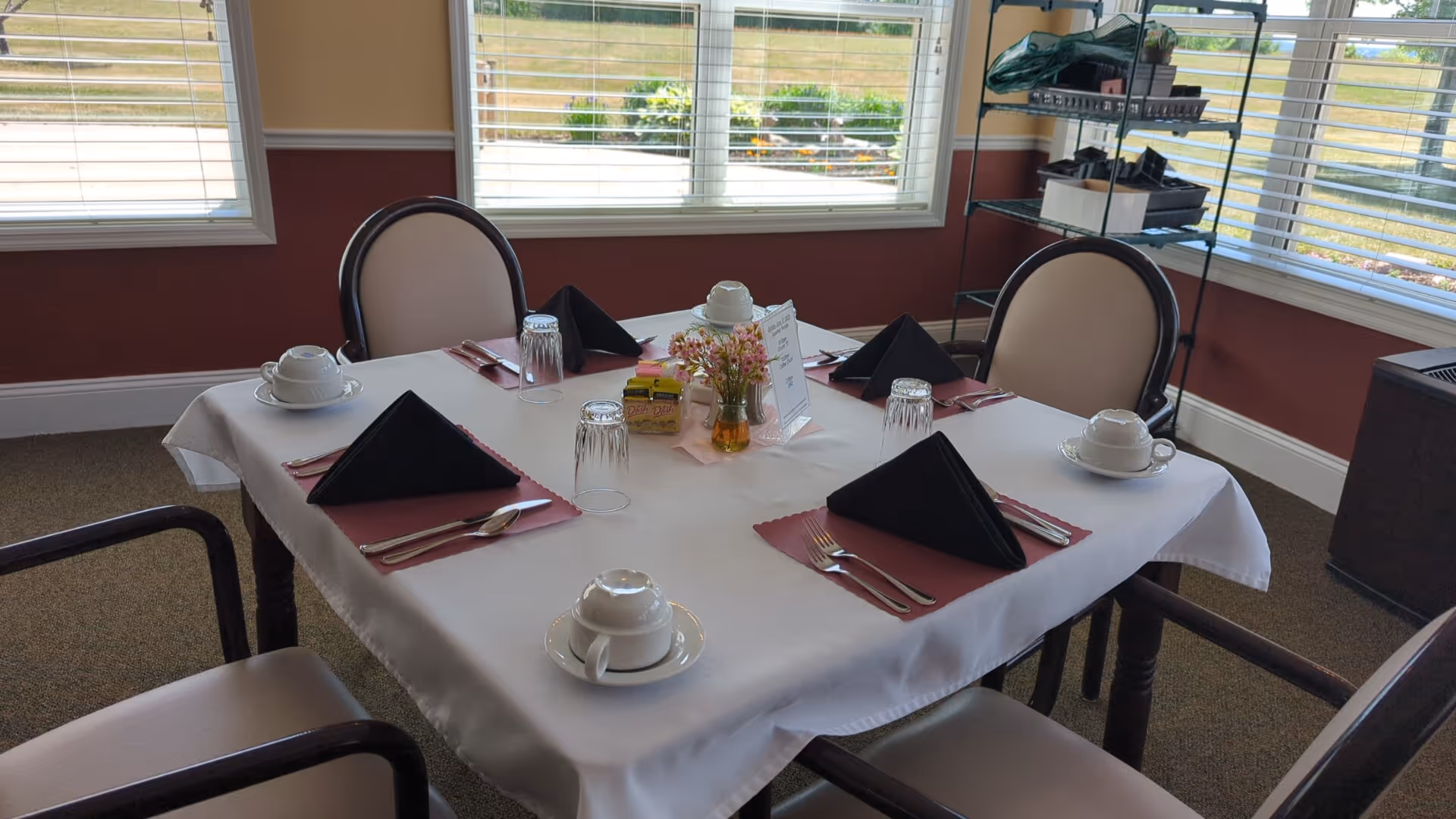 A dining table set for four with white tablecloth, black folded napkins, cups and saucers, glasses, and silverware. The table is near windows with blinds, and there is a small flower arrangement and condiments in the center. Four chairs with light-colored cushions surround the table.