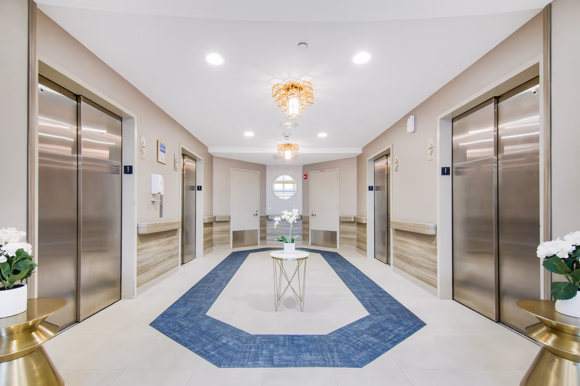 A bright and clean elevator lobby area with four stainless steel elevators, two on each side. The floor features a blue and beige geometric rug, and a small round table with a white orchid plant is centered in the middle. The walls are painted light beige with wood paneling halfway up. Two modern ceiling light fixtures hang from the white ceiling. There are two closed doors at the end of the hallway with a round window above them. Two small gold side tables with white flower pots are placed near the elevators.
