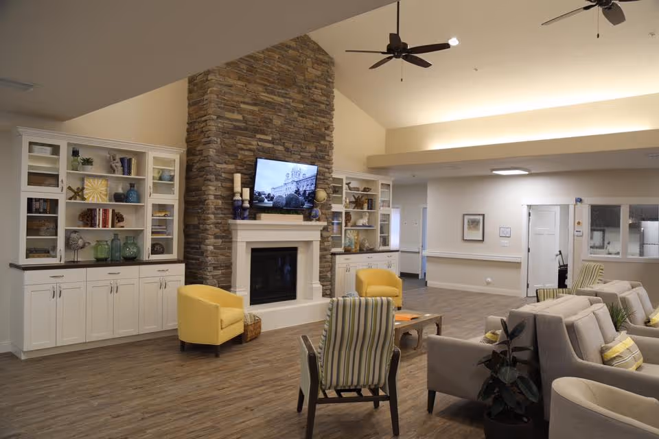 Spacious assisted living common room with a stone fireplace, wall-mounted TV, built-in cabinets and multiple seating areas.