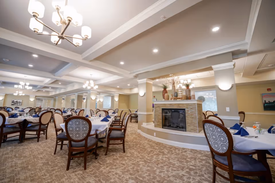 A spacious dining room with multiple round tables covered with white tablecloths and set with blue napkins, glasses, and small floral centerpieces. The room features beige walls, carpeted floors, elegant chandeliers, and a central decorative fireplace with vases and plants on top.