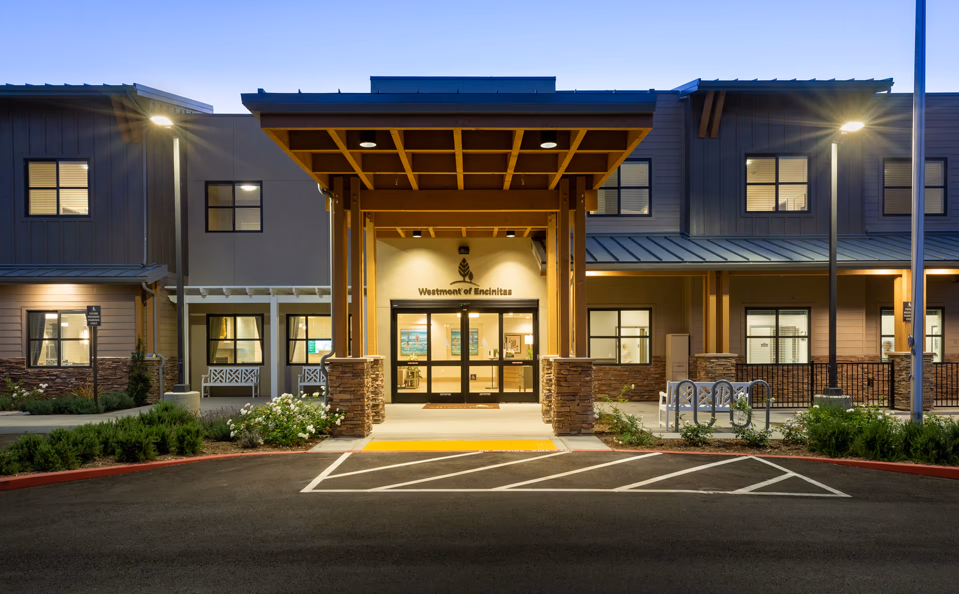 Exterior front entrance of Westmont of Encinitas facility at dusk, featuring a covered entryway with stone pillars, illuminated by outdoor lights, with windows and landscaping visible on either side.