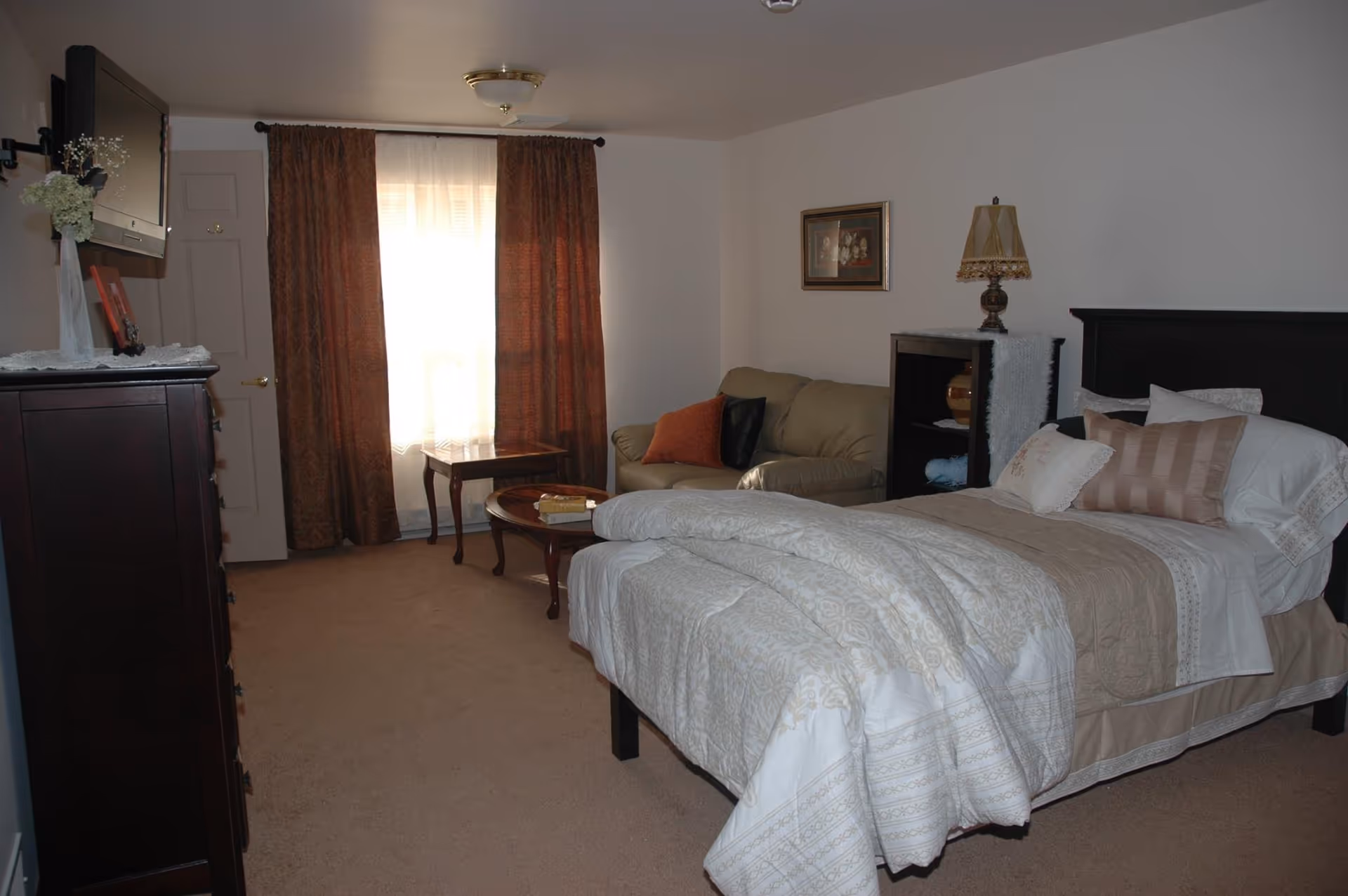 A bedroom with a neatly made bed featuring beige and white bedding, a beige loveseat with orange and black pillows, a wooden coffee table and side table, a dark wooden dresser with a vase of flowers and picture frames, a wall-mounted TV, brown curtains covering a window, and a ceiling light fixture.