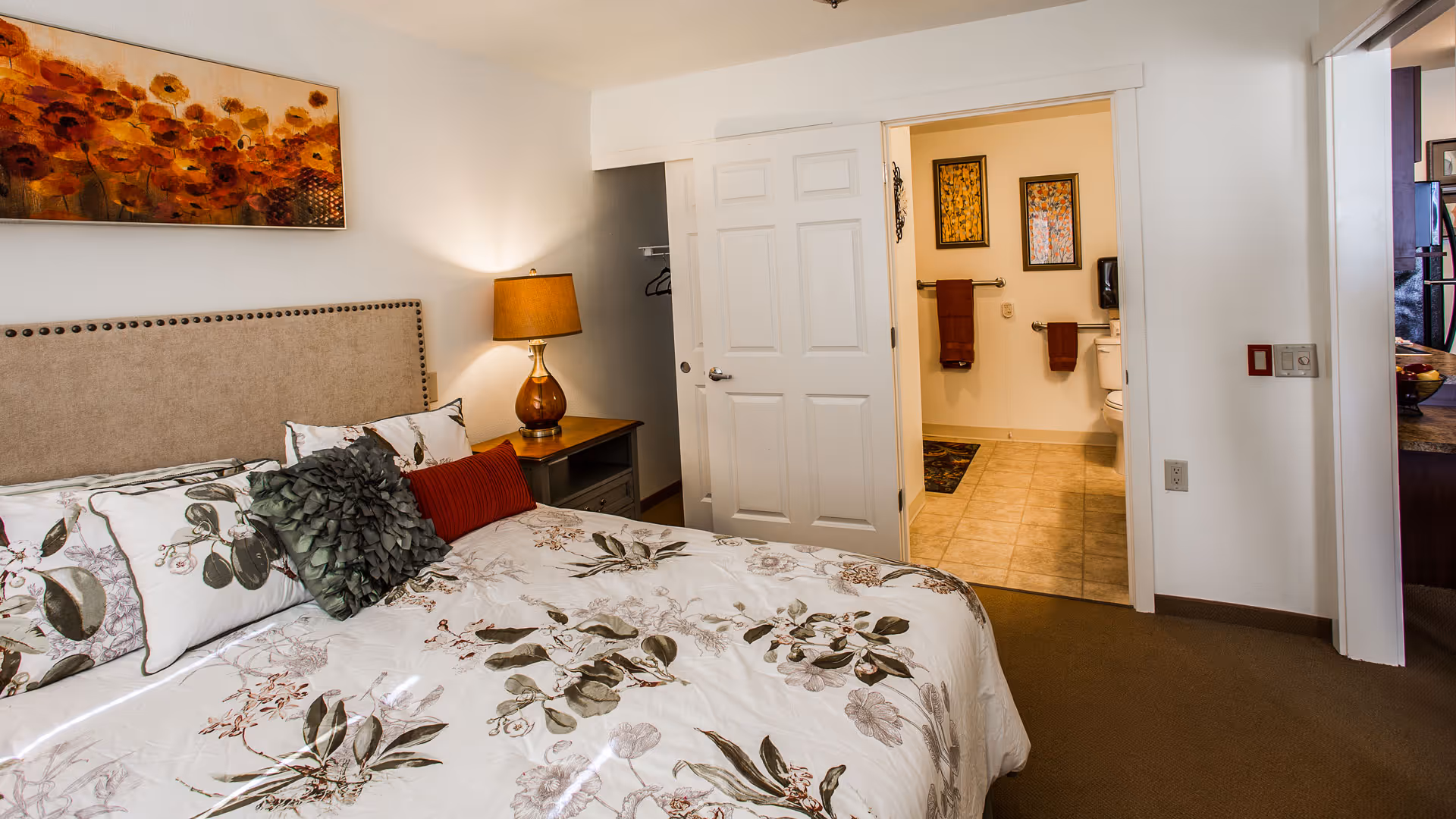 A cozy bedroom with a floral patterned bedspread, decorative pillows, a beige upholstered headboard, and a bedside table with a lamp. The room has white walls with a painting of orange flowers above the bed. An open door reveals a bathroom with tiled flooring, two framed pictures, and towels hanging on the wall.