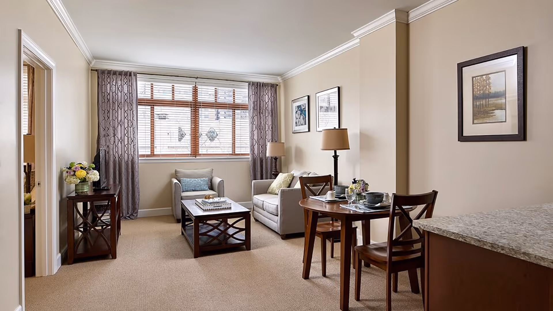 A cozy living room area with a beige sofa, an armchair, a wooden coffee table, and a small dining table set for two. The room has beige walls, a large window with wooden blinds and purple curtains, two table lamps, framed artwork on the walls, and a side table with a flower arrangement.