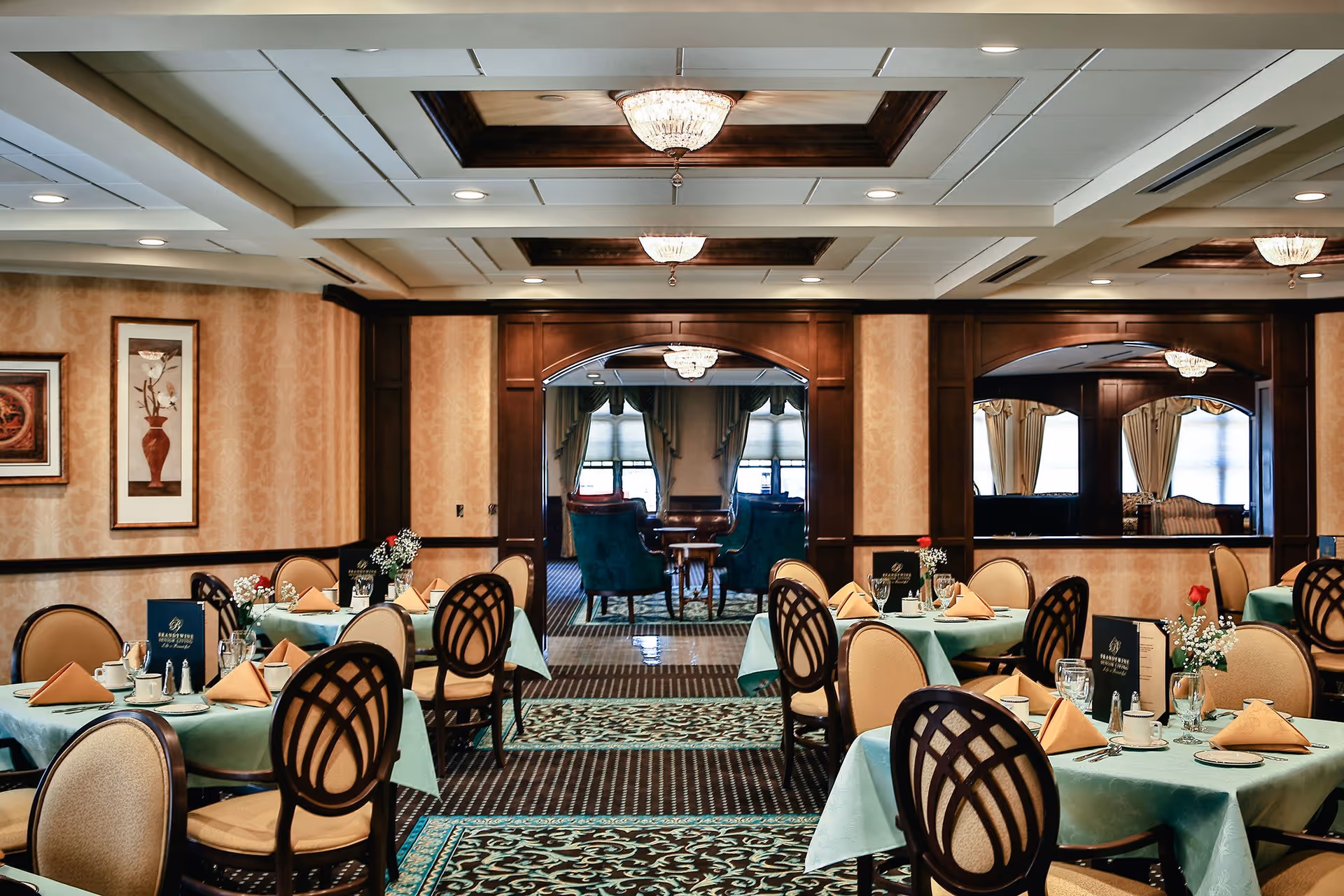 Elegant dining room with tables covered in light blue tablecloths, each set with beige napkins, glassware, and small floral centerpieces. The room features patterned carpet, beige wallpaper, framed artwork, and dark wood paneling with arched openings leading to a lounge area with green upholstered chairs and large windows with curtains.