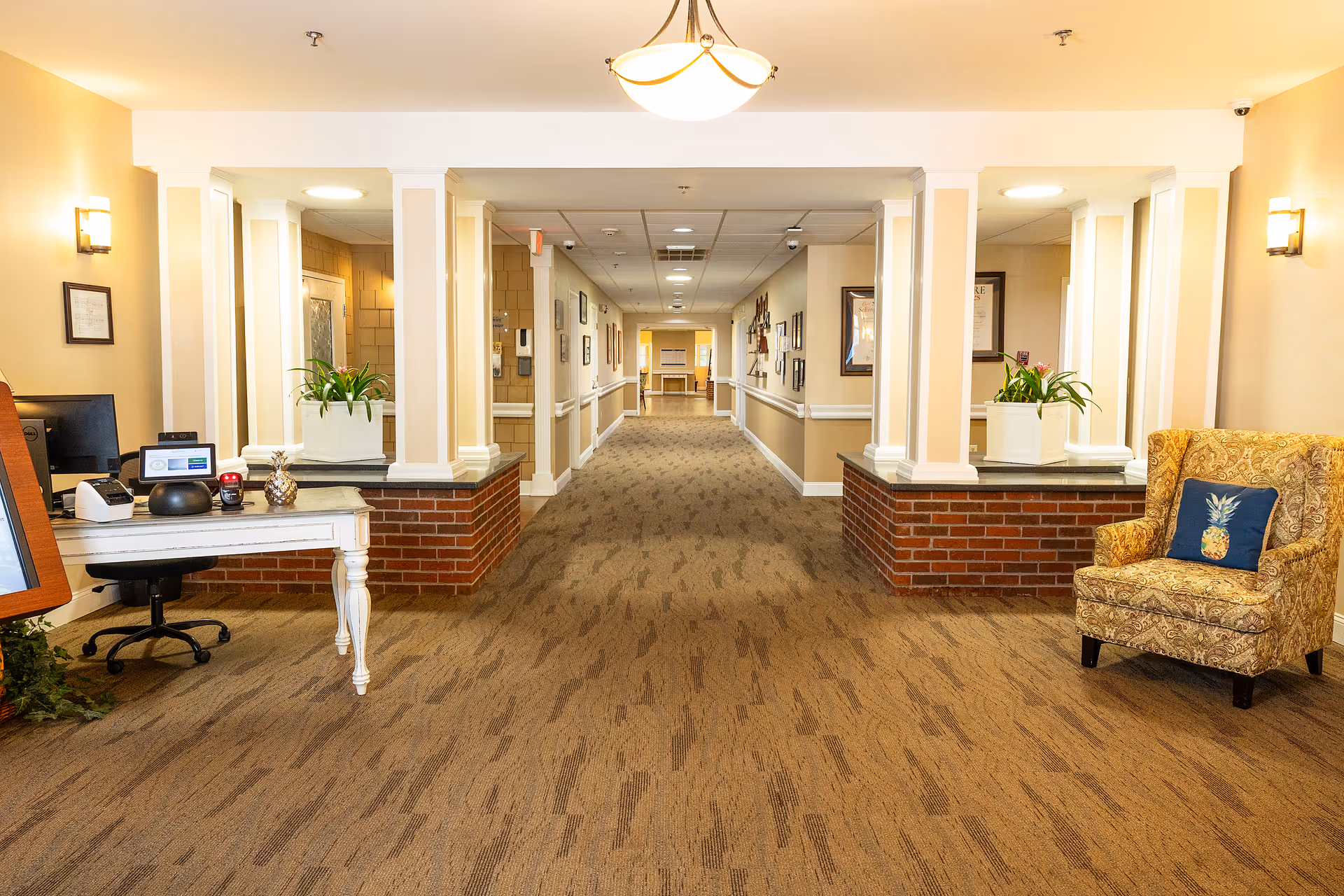A well-lit hallway in a senior living facility with beige walls and carpeted floor. On the left side, there is a white desk with a computer and decorative items, and on the right side, there is a patterned armchair with a pineapple-themed pillow. The hallway extends into the distance with framed pictures on the walls and plants placed on brick half-walls near the entrance.