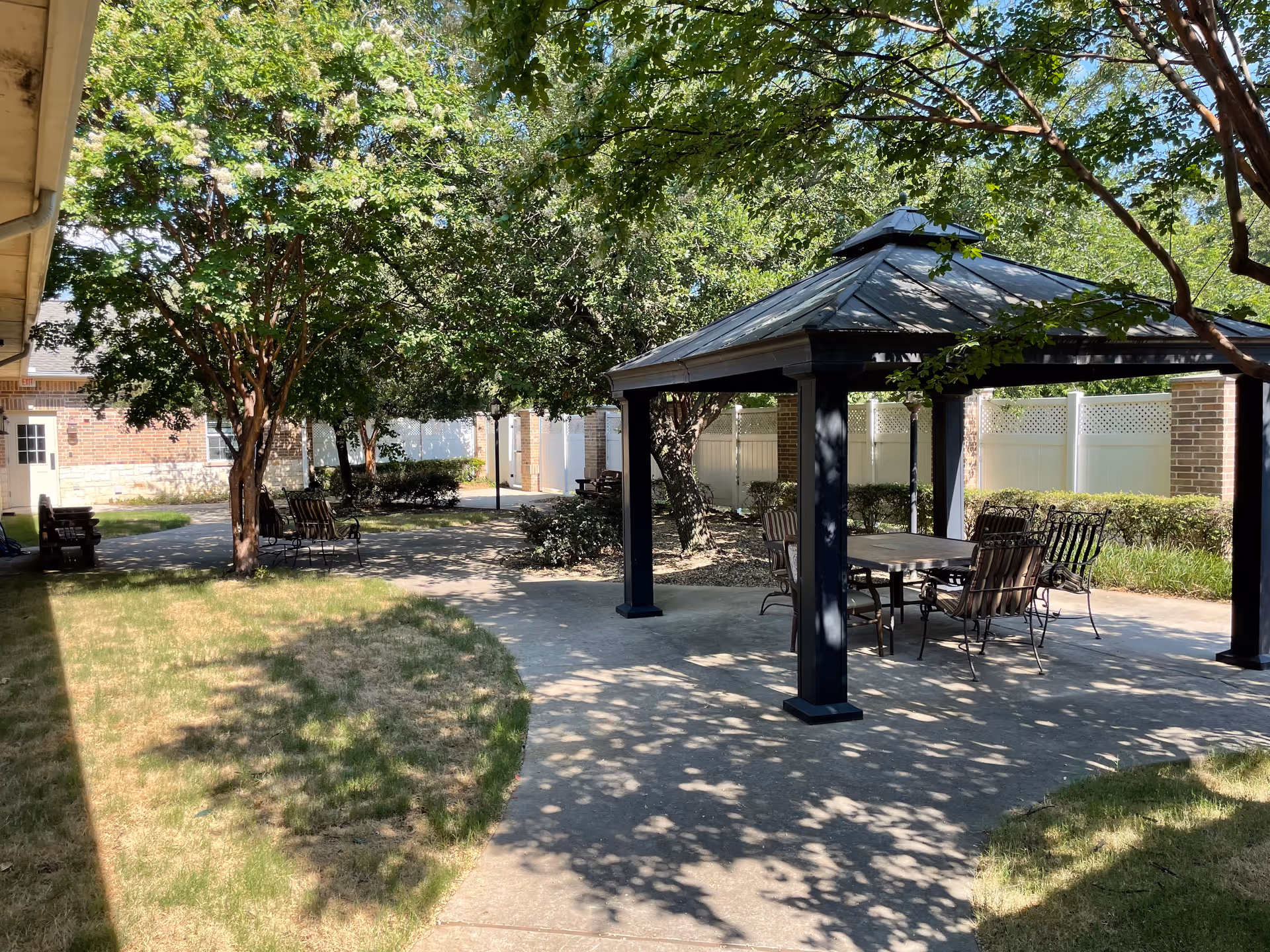Outdoor patio area at Magnolia Assisted Living and Memory Care featuring a gazebo with a table and chairs underneath, surrounded by trees and greenery, with a paved walkway and a building in the background.