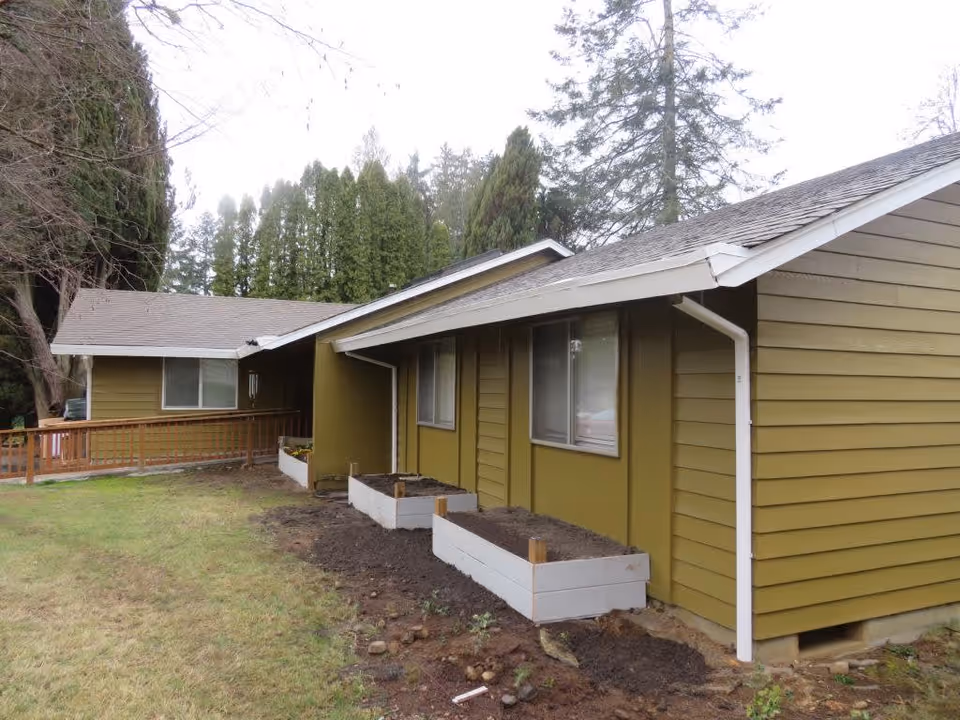 Exterior view of a single-story building with olive green siding and a gray shingled roof. There are three rectangular raised garden beds filled with soil along the side of the building. The area around the building has grass and trees in the background under an overcast sky.