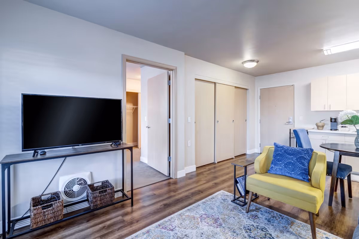 A modern living room area with a flat-screen TV on a black metal stand, two wicker baskets underneath, a yellow armchair with a blue cushion, a small side table, a round dining table with blue chairs, and light wood flooring. The room has white walls, a ceiling light, and an open door leading to a closet and another room.