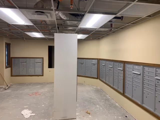 An indoor mailroom area with multiple gray mailboxes mounted on beige walls. The floor is unfinished concrete with some debris and a white cloth on the ground. The ceiling is partially open, exposing ducts and pipes, with fluorescent lights providing illumination.