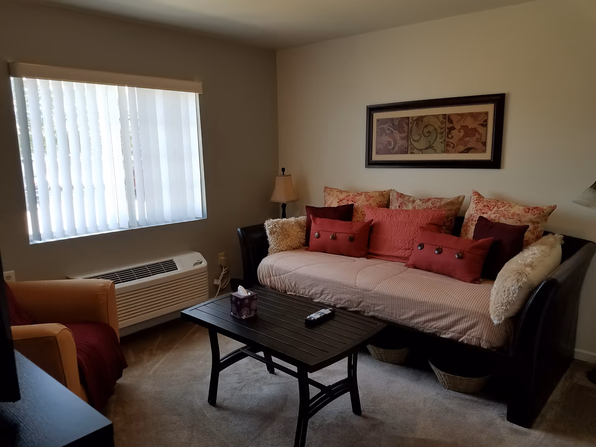 Cozy living room with a daybed covered in patterned and red pillows, a coffee table, armchair, lamp, and a window with vertical blinds.