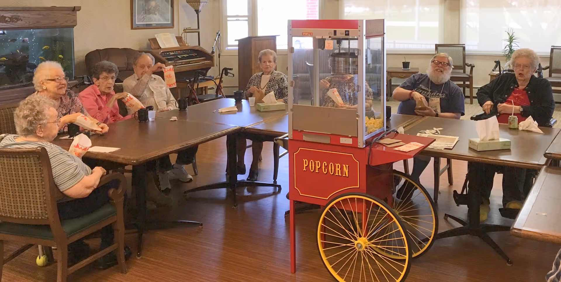 A group of seniors seated around tables in a common room enjoying popcorn from a red popcorn cart.
