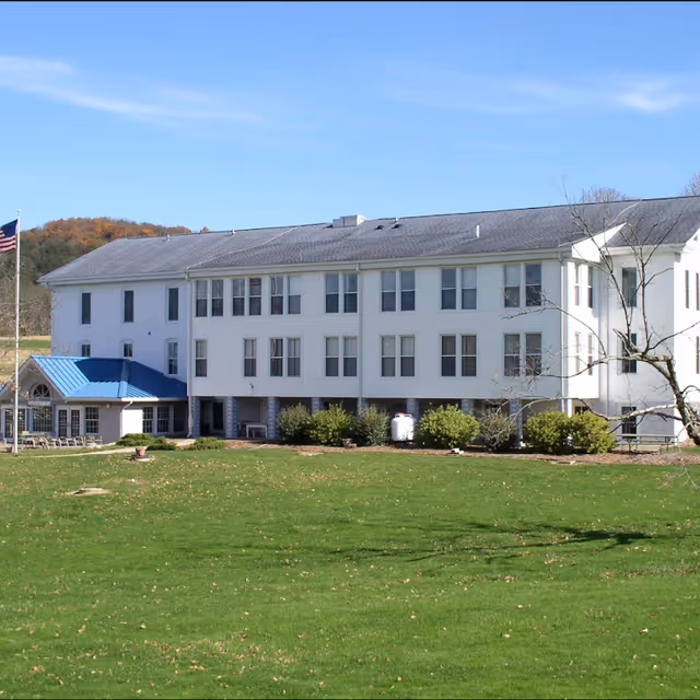 Exterior view of a large white multi-story building with many windows, a blue roofed entrance, an American flag on a flagpole, and a well-maintained green lawn in front under a clear blue sky.