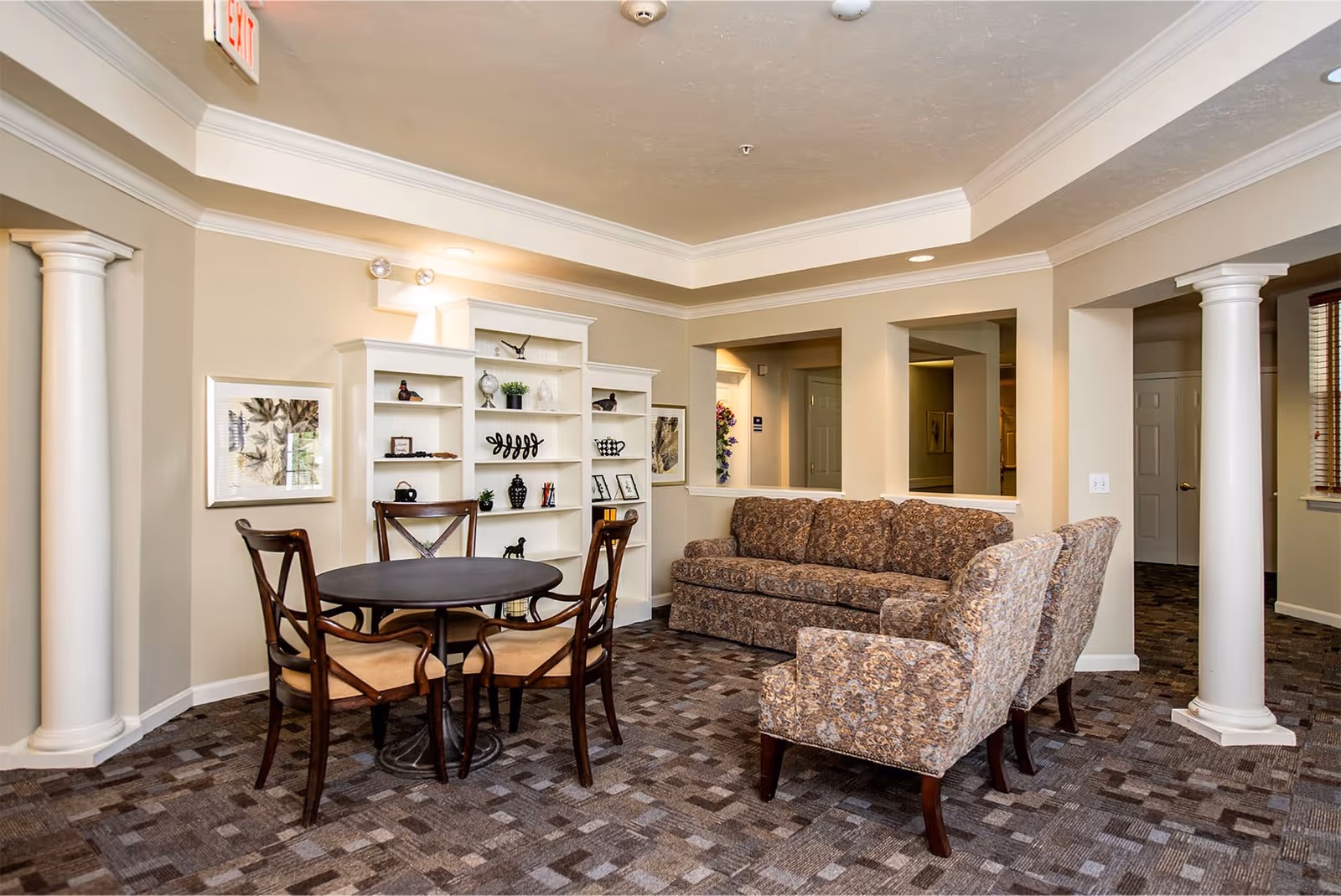 Well-lit common living room with a round table and chairs, upholstered sofa and armchairs, and built-in shelving.