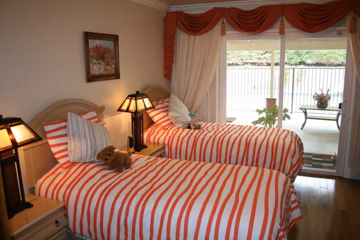 A bedroom with two twin beds featuring red and white striped bedding, each with a pillow and a stuffed animal. There are two bedside tables with lamps, a framed painting on the wall, and a sliding glass door leading to an outdoor patio area with a bench and plants.