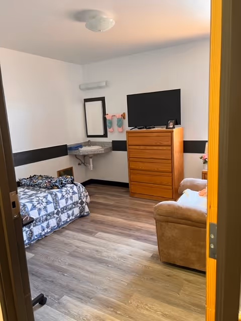 Small bedroom with a single bed, a dresser topped by a TV, a wall-mounted sink, and an upholstered chair.