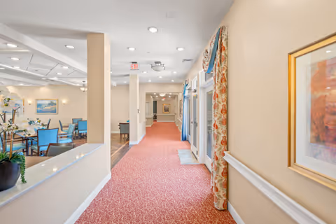 Carpeted interior hallway in a senior living facility with dining seating visible to the left and decorative curtains along the windows on the right.