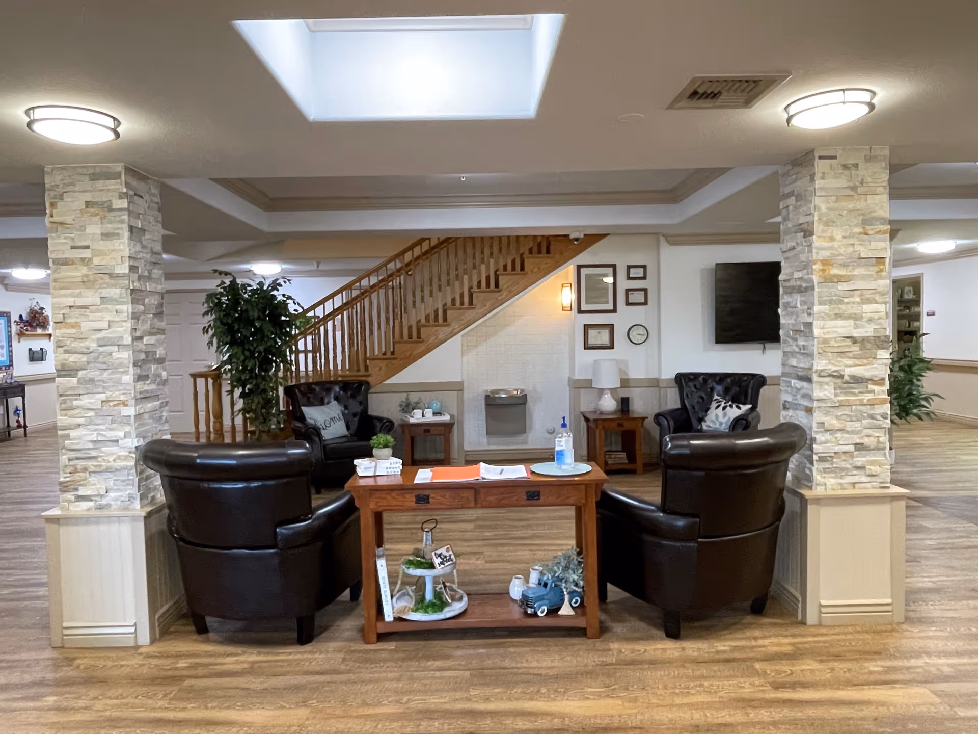 A cozy seating area in an assisted living facility featuring four dark leather armchairs arranged around a wooden table with decorative items. The space includes two stone pillars, a staircase in the background, a water fountain, a wall-mounted TV, and soft overhead lighting.