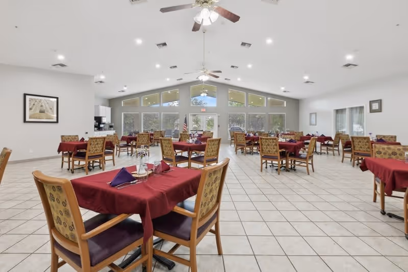A spacious dining room with multiple tables covered in burgundy tablecloths and set with napkins and glasses. The room has tiled floors, large windows letting in natural light, ceiling fans, and light-colored walls with minimal decorations.