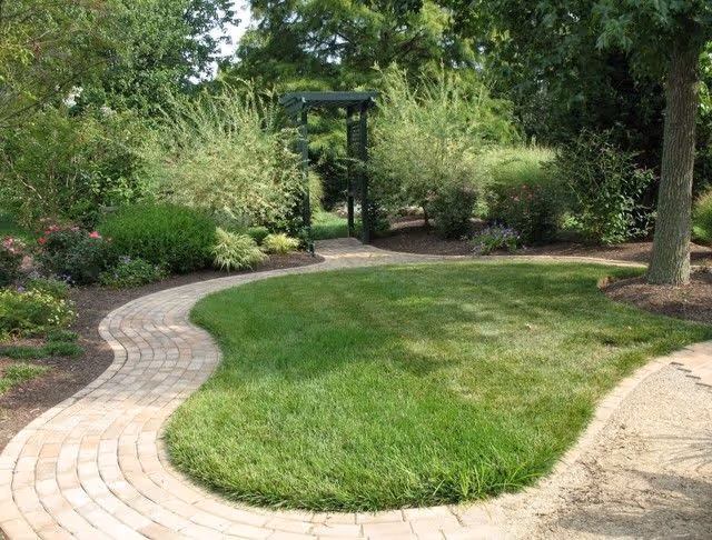 Curved brick pathway winds through a landscaped garden with a central lawn and a wooden arbor in the background.