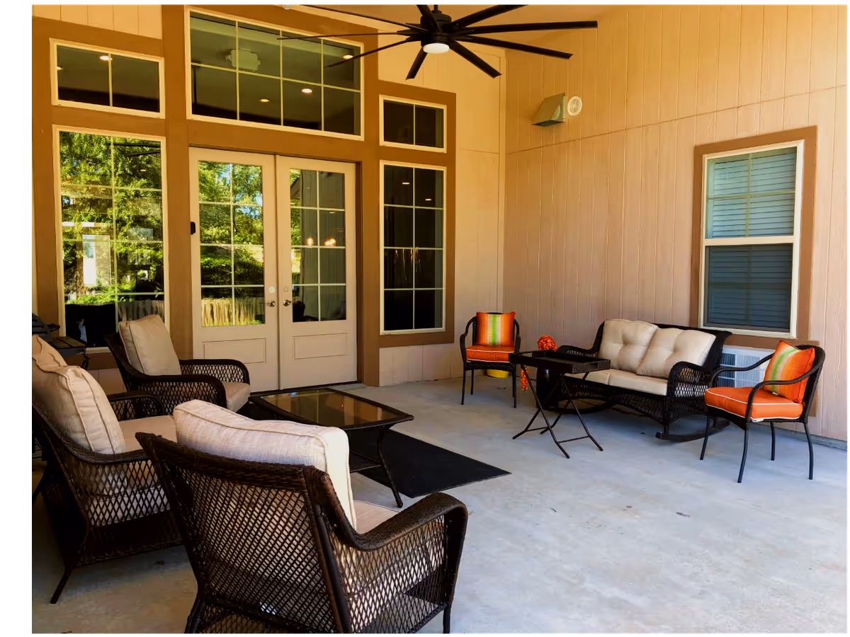 Covered outdoor patio area with cushioned wicker chairs, a cushioned wicker loveseat, two orange cushioned chairs, a glass-top coffee table, and a small black folding table. The patio has beige walls, a ceiling fan, and large windows and double doors leading inside.
