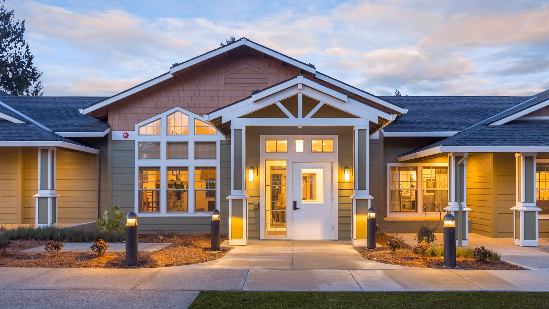 Front entrance of a single-story senior living building with lit windows and walkway lights at dusk.
