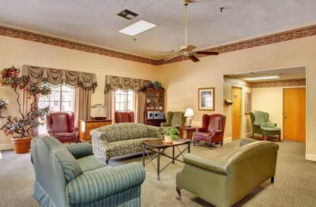 A cozy living room area in a nursing and rehabilitation facility featuring multiple upholstered armchairs and sofas arranged around a coffee table with a small plant. The room has beige walls, carpeted floor, two windows with valance curtains, a ceiling fan, framed artwork, and a potted plant in the corner. There is an open doorway leading to another room with additional seating.