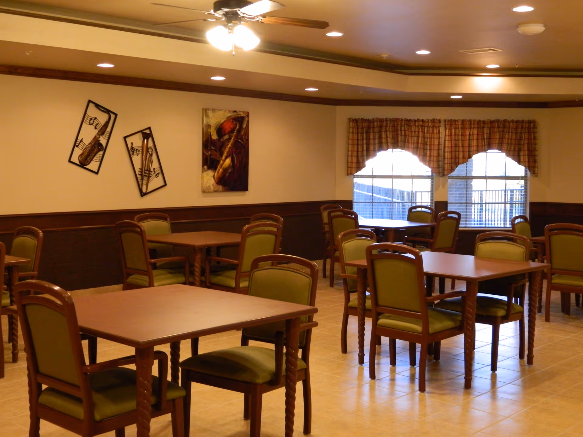 A dining room with several wooden tables and green cushioned chairs. The room has beige walls with dark wood paneling, three pieces of musical-themed wall art, two windows with plaid valances, and a ceiling fan with lights.