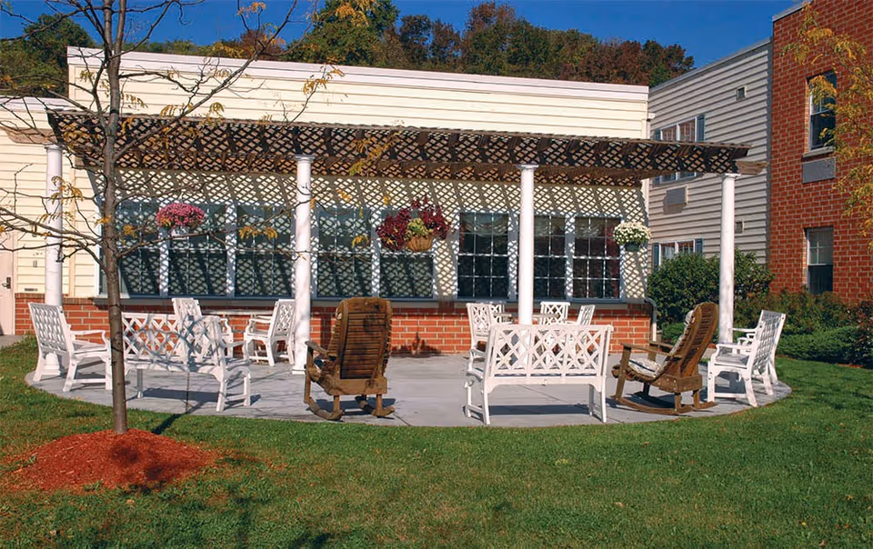 Outdoor patio area at a senior living facility with white and wooden chairs arranged in a circle on a concrete surface. The patio is shaded by a lattice pergola attached to a building with large windows. There are hanging flower baskets and a small tree with mulch in the foreground on a grassy lawn.