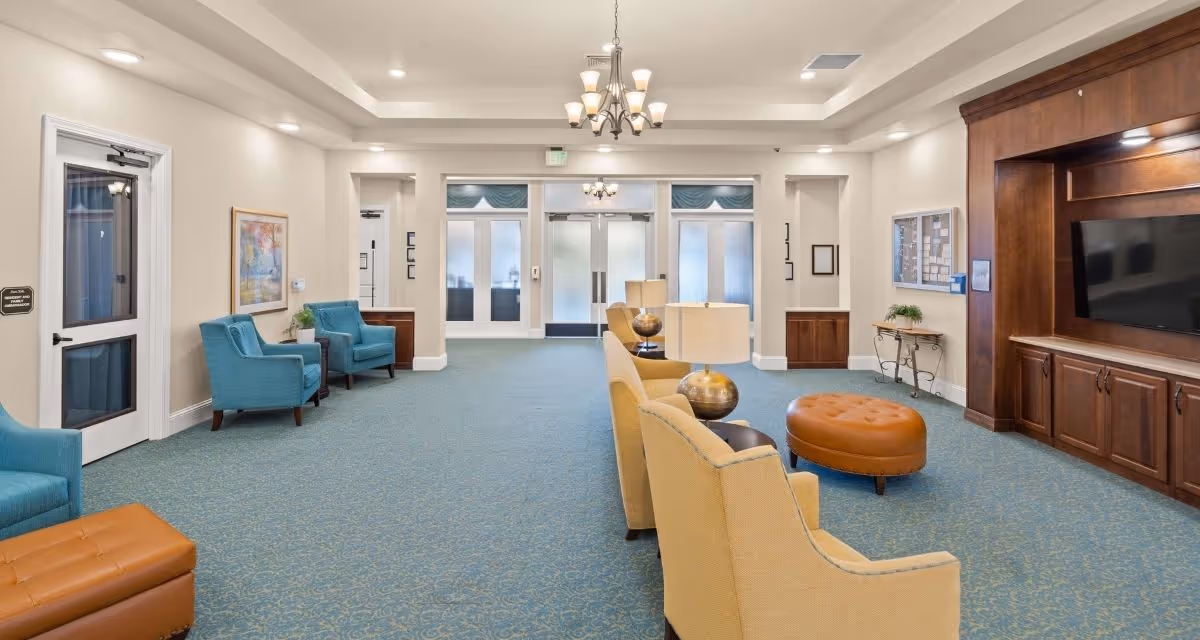 A spacious and well-lit common area in a senior living facility featuring blue carpet with a subtle pattern, beige walls, and a tray ceiling with recessed lighting and a chandelier. The room is furnished with yellow armchairs, blue upholstered chairs, a brown leather ottoman, and side tables with lamps. A large wooden entertainment center with a flat-screen TV is on the right wall. There are double glass doors at the far end of the room, and framed artwork and plants decorate the space.