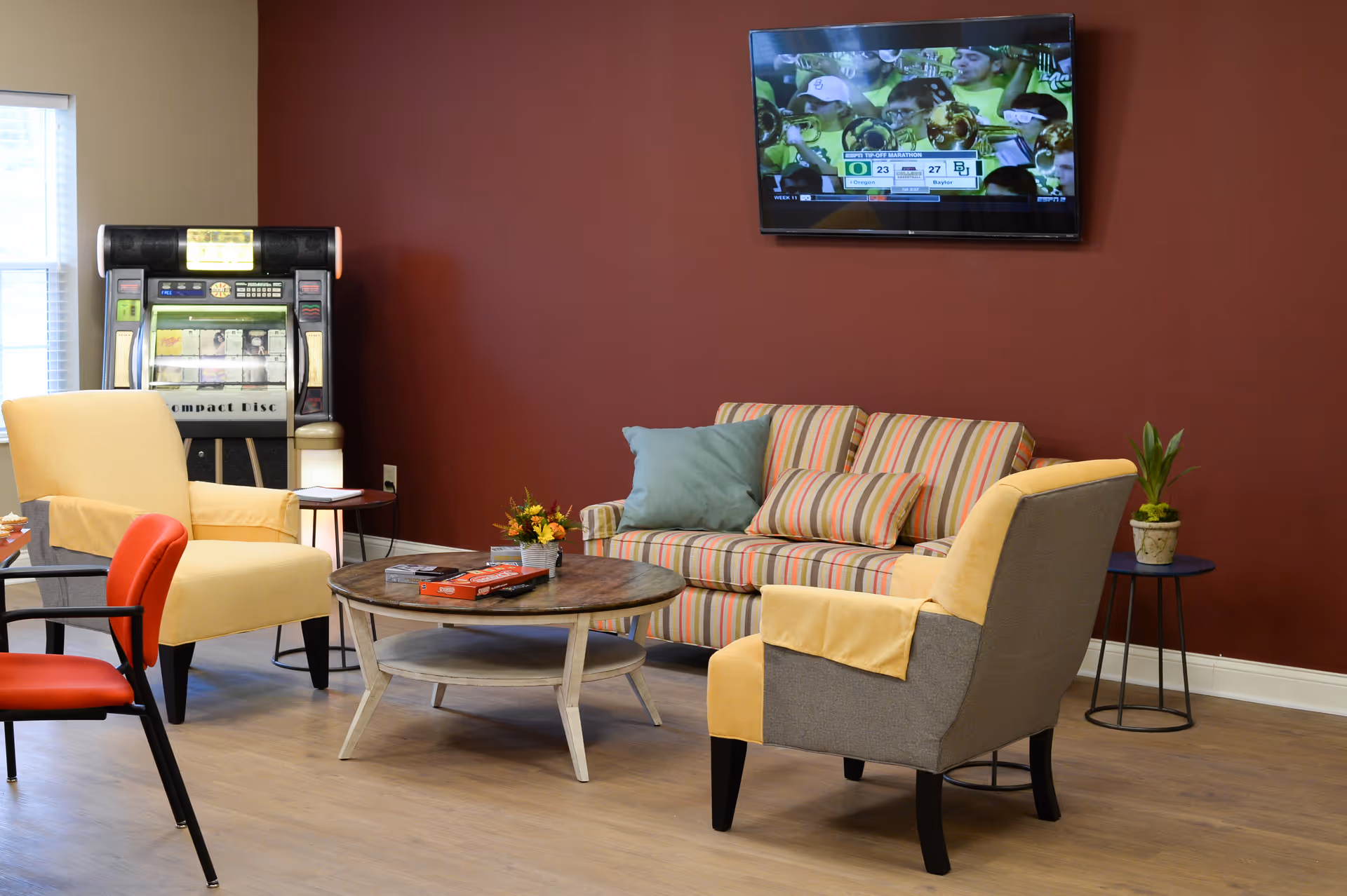 A cozy living room area with a striped sofa adorned with two pillows, two yellow armchairs with gray sides, a round wooden coffee table with board games and a remote control, a small side table with a plant, a jukebox near a window, and a wall-mounted TV showing a sports game.
