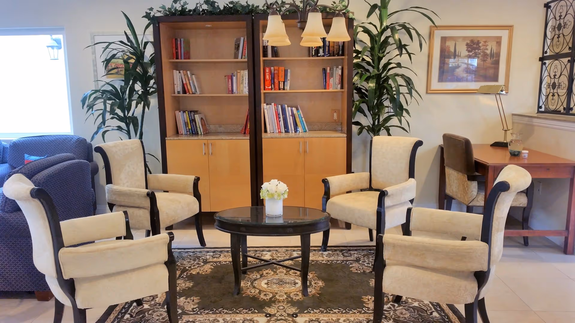 A cozy sitting area with four beige upholstered chairs arranged around a round glass-top coffee table with a small white flower arrangement. Behind the chairs are two wooden bookshelves filled with books and two tall green plants on either side. To the right, there is a wooden desk with two chairs, a lamp, and a framed picture on the wall. A patterned rug covers the floor beneath the seating area.