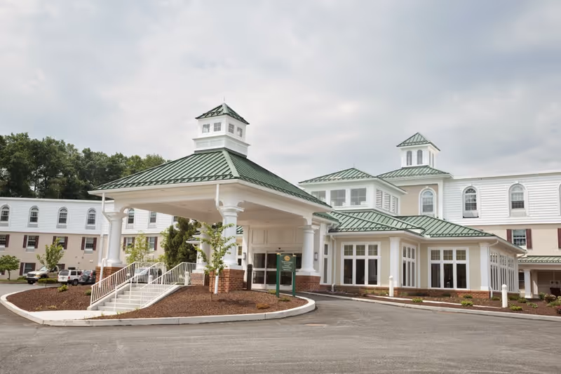Exterior view of a large retirement community building with white walls and green metal roofs, featuring a covered entrance with columns and a circular driveway. There are some small trees and landscaping around the entrance, and several parked cars are visible on the left side.