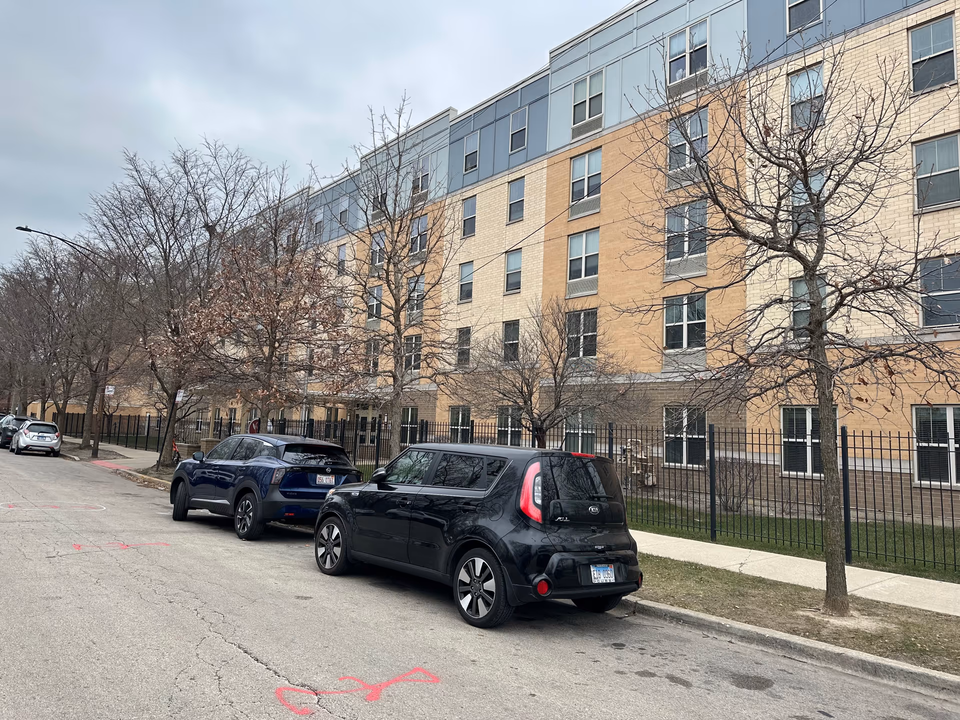 Street view of a multi-story residential building with beige and blue exterior walls, leafless trees along the sidewalk, and several parked cars on the street in front of the building under an overcast sky.