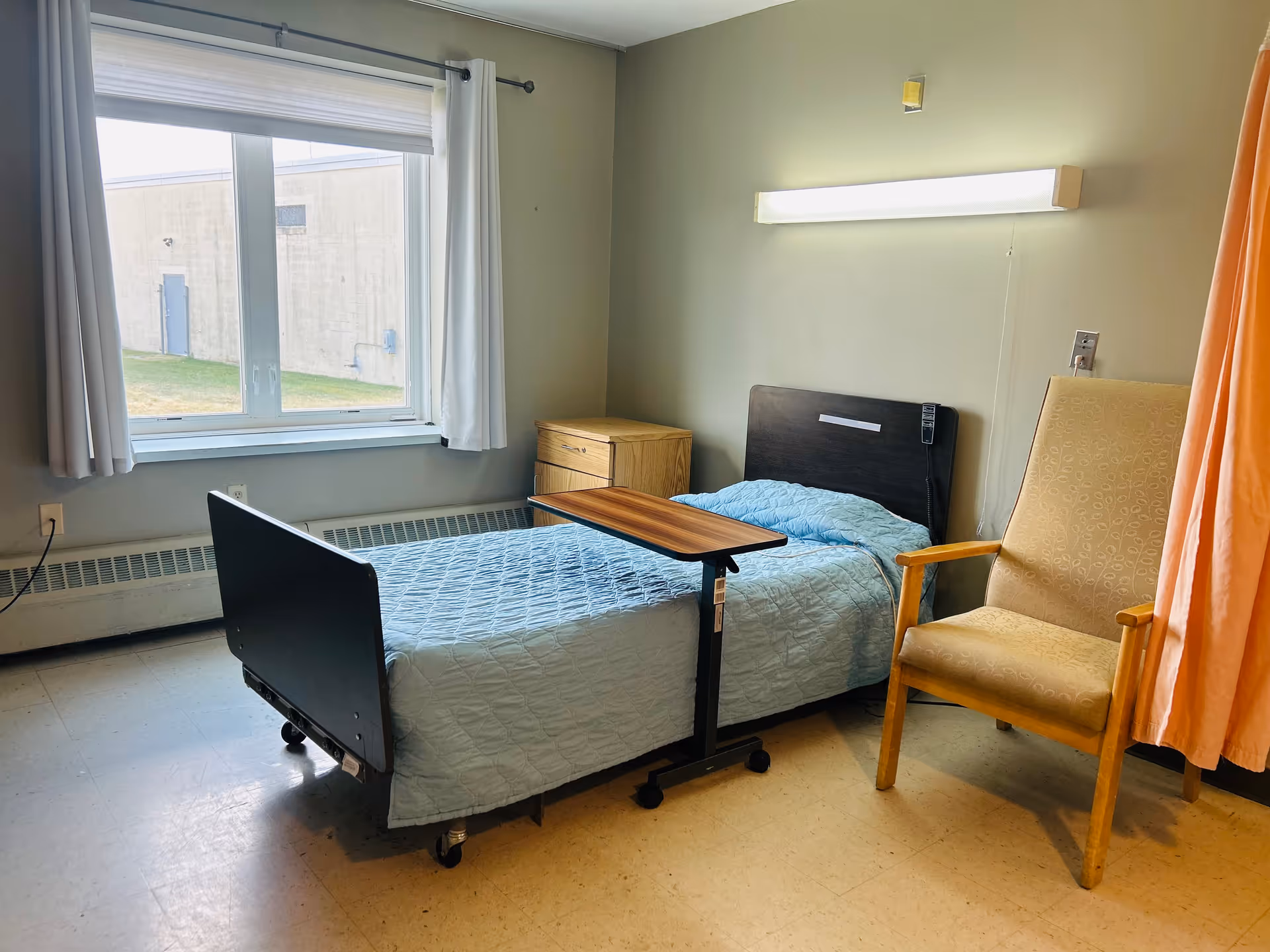 A simple room with a single hospital-style bed covered with a light blue quilt, a wooden bedside table, a beige armchair, a window with white curtains, and a wall-mounted light above the bed. There is a small overbed table positioned over the bed.