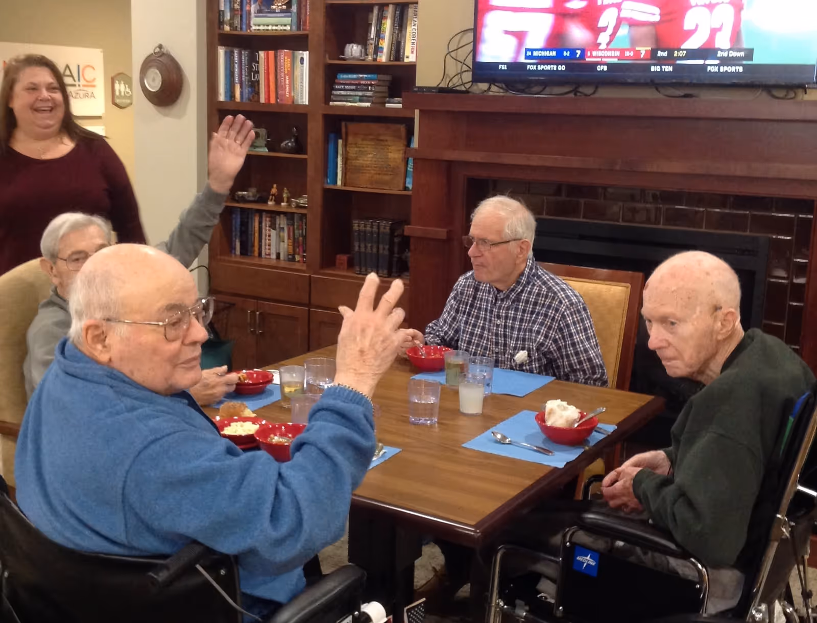 Four elderly men sitting around a wooden table eating from red bowls with drinks, while a woman stands nearby smiling. The setting appears to be a cozy common area with bookshelves, a fireplace, and a television mounted above it.