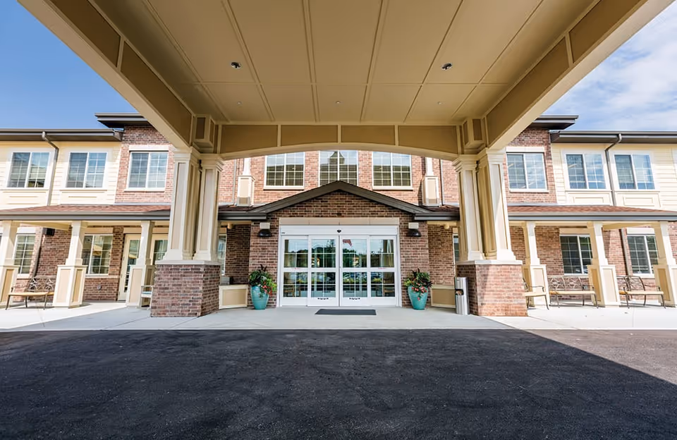 Entrance of Dougherty Ferry Assisted Living and Memory Care facility showing a covered drop-off area with large pillars, glass double doors, brick and beige exterior walls, and potted plants on either side of the entrance.