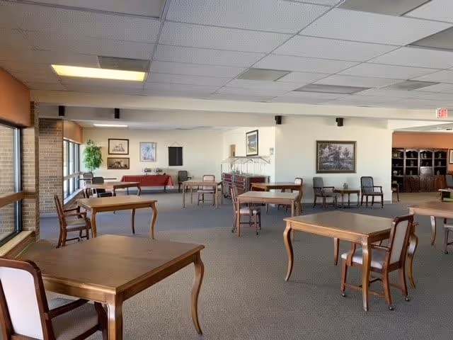 A spacious room with several wooden tables and chairs arranged throughout. The room has large windows on one side letting in natural light, framed pictures on the walls, and a carpeted floor. There is a buffet or serving area along one wall and a few chairs lined up against the walls.