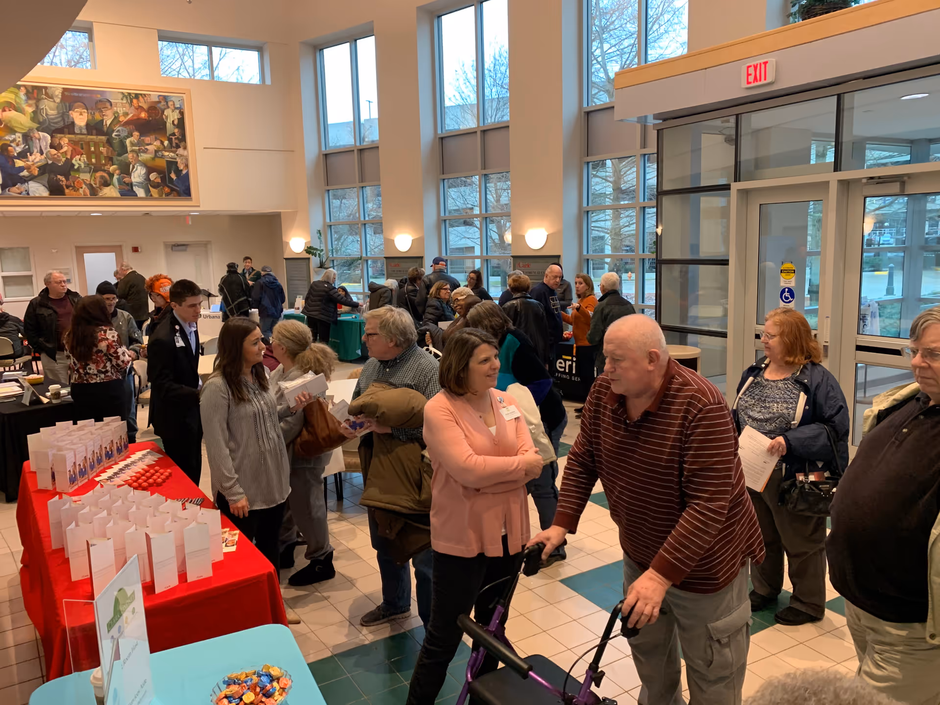 A busy indoor lobby area with many people interacting and standing in line near tables covered with red and green tablecloths. The space has large windows letting in natural light, a high ceiling, and a large painting on the wall. An elderly man with a walker is in the foreground, and several other adults are engaged in conversation or waiting.