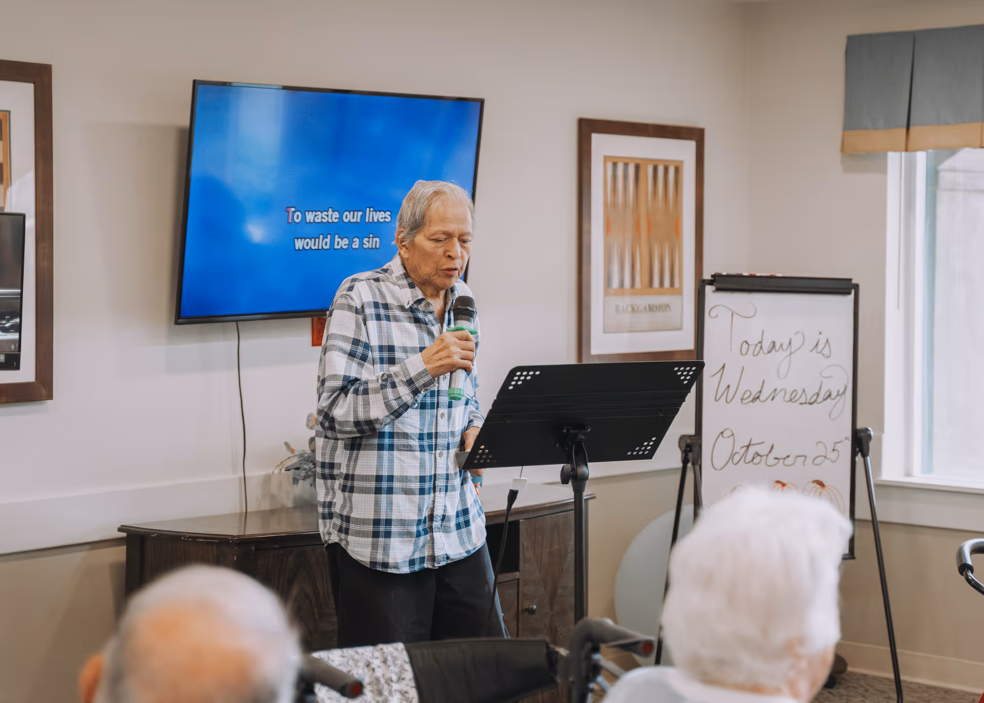 An elderly man in a plaid shirt is singing into a microphone in a room with a TV screen displaying karaoke lyrics. In the foreground, two elderly people are seated, watching him. A whiteboard in the background shows the date as Wednesday, October 25.