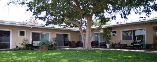 Outdoor courtyard area of a senior living facility with a large tree in the center, surrounded by a curved building with multiple sliding glass doors and windows. There are patio chairs and potted plants along the building's exterior, and a well-maintained green lawn in the foreground.