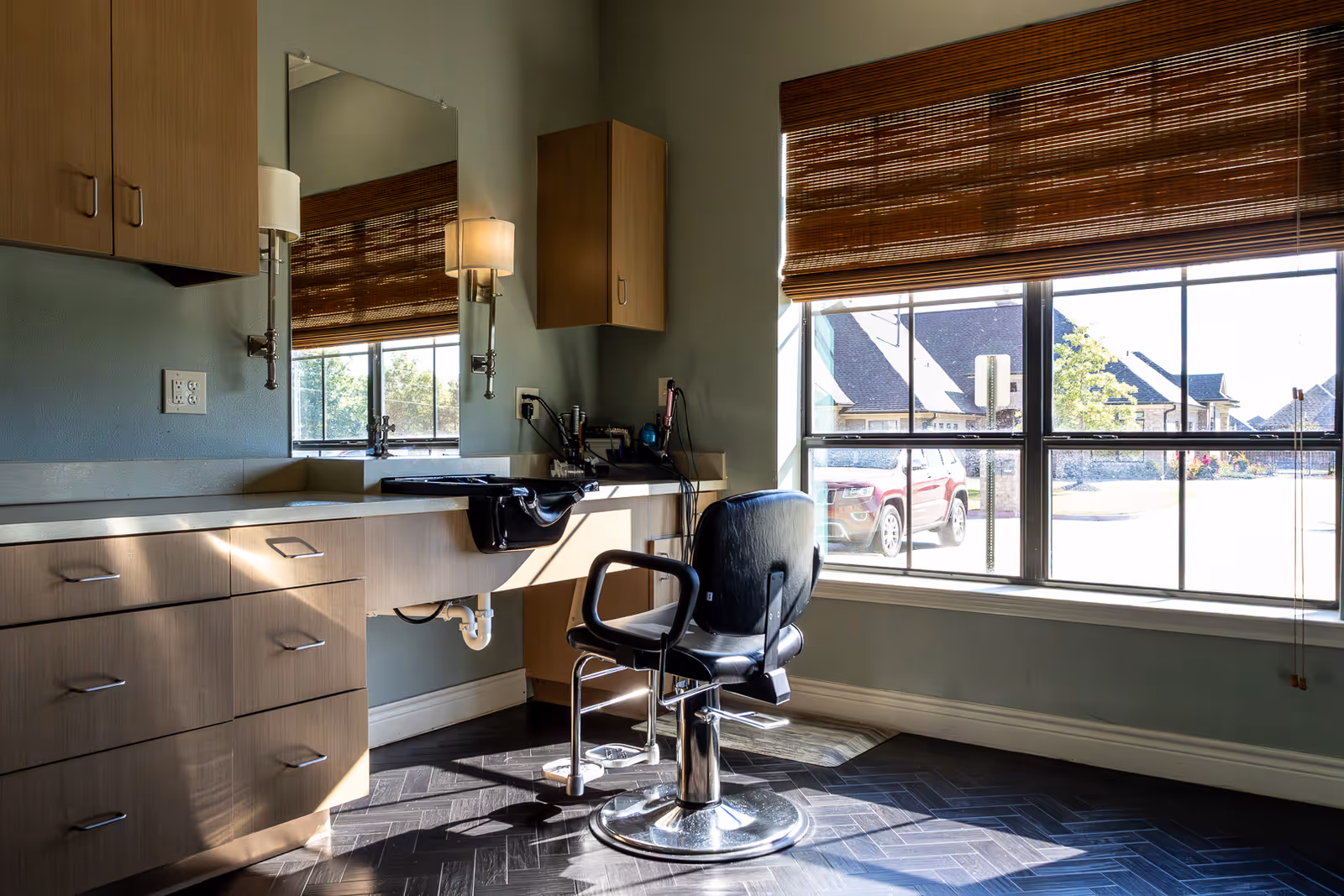A well-lit room with a large window covered by a bamboo blind, featuring a black salon chair in front of a counter with a built-in sink and mirror. The counter has multiple drawers and cabinets, and there are wall-mounted cabinets and a wall lamp beside the mirror. Outside the window, a red vehicle and houses are visible.