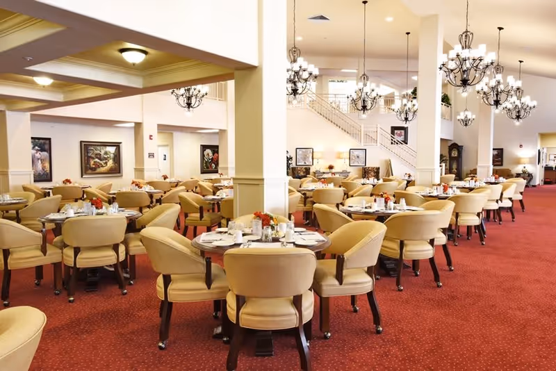 A spacious dining room with multiple round tables set with plates, glasses, and silverware. Each table is surrounded by beige upholstered chairs. The room has red carpeting, white walls, and several chandeliers hanging from the ceiling. There are framed paintings on the walls and a staircase visible in the background.