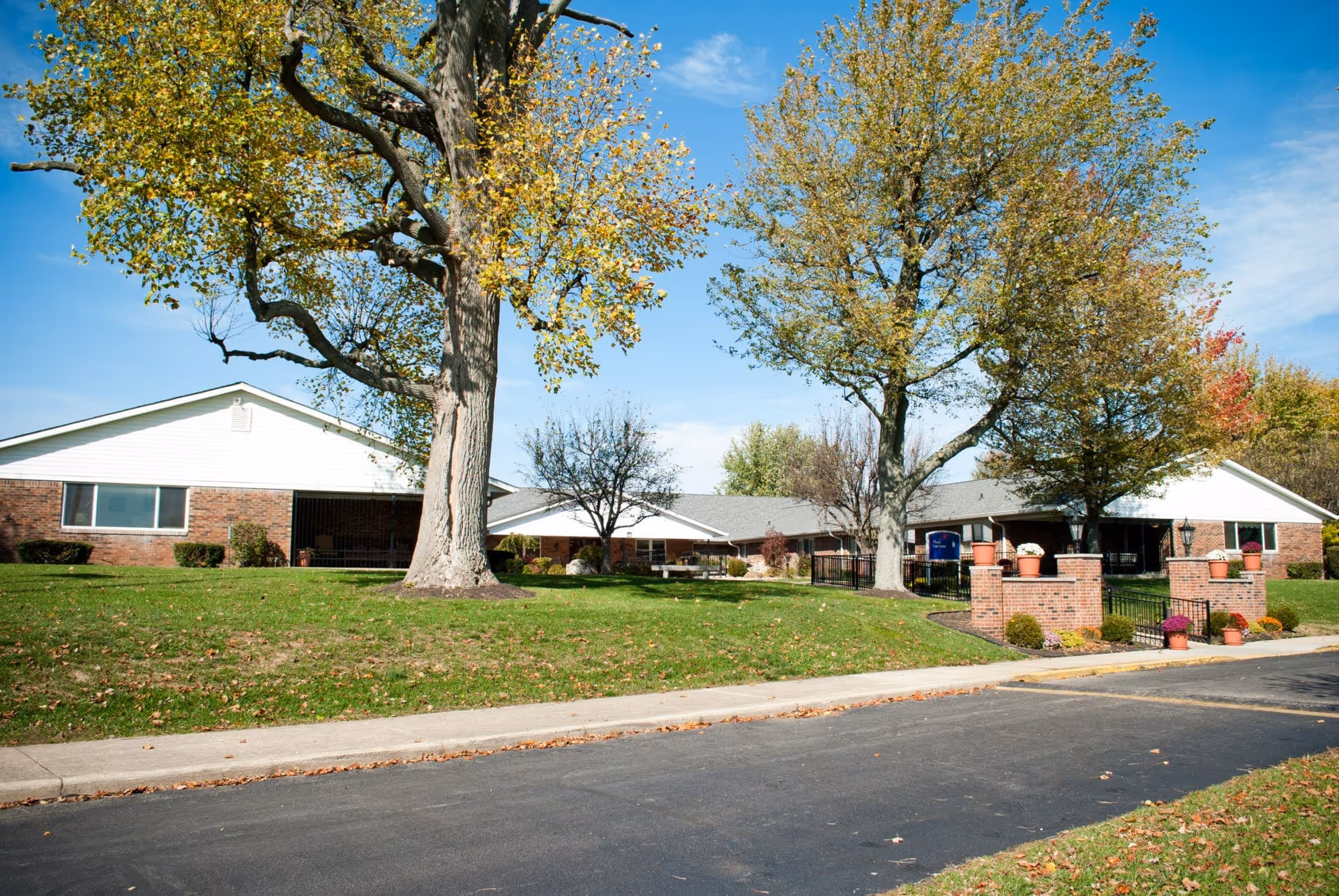 Exterior view of Arbor Grove Village facility with a brick building, large trees with autumn foliage, green lawn, and a paved road in front. There are potted plants on brick pillars near the entrance.