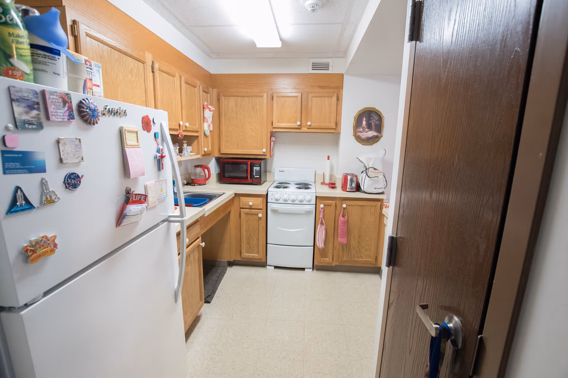 Small kitchen with oak cabinets, a white refrigerator covered in magnets, a stove, microwave, sink, and countertop appliances.