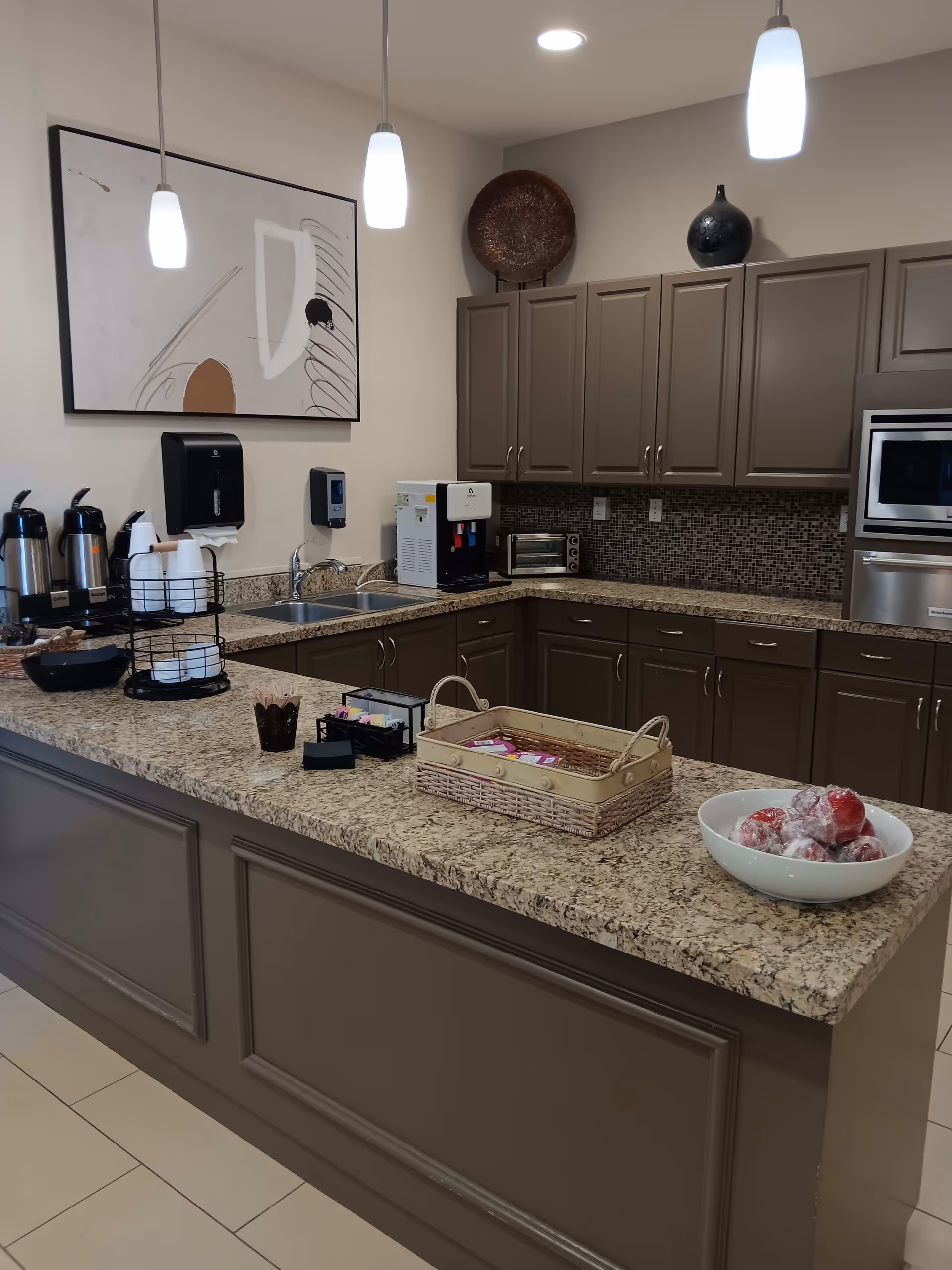 A modern kitchen area with granite countertops and brown cabinets. The countertop has a basket with packets, a bowl of wrapped apples, and coffee supplies including dispensers and cups. Above the counter hang three pendant lights. On the wall, there is abstract artwork and decorative items on top of the cabinets.
