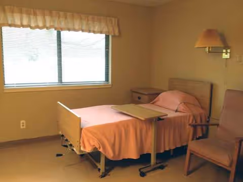 Empty patient bedroom in a senior living facility with pink bedspread, window, and chair.